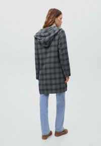 Kvinna som vänder bort, klädd i en grå rutig hooded kappa, ljusblå jeans och bruna skor mot en enfärgad grå bakgrund.