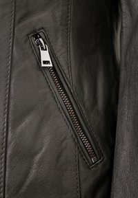 Schwarze Lederjacke mit sichtbar seitlichem Reißverschluss; der Reißverschluss-Zip ist silberfarben; glatte Textur mit dezentem Narbenmuster.