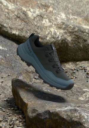 Musta ja sininen Mammutin retkeilykenkä, jossa on kestävä pohja, roikkuen kivikkoisen maaston yläpuolella, jossa on pieniä kiviä ja lohkareita.