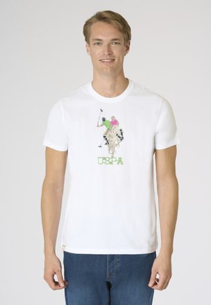 Uomo che indossa una maglietta bianca con giocatori di polo colorati e testo verde "USPA", abbinata a jeans blu, in piedi davanti a uno sfondo semplice.