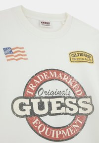 Witte katoenen T-shirt met een groot, verweerd rood en zwart cirkelvormig logo met "GUESS" en "Originals." Inclusief een kleine vlagafbeelding en een patch.