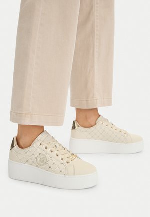 Beżowe sneakersy na platformie z perforowanym wzorem, złotymi oczkami i metalicznym detalem na pięcie, noszone z jasnobeżowymi spodniami.