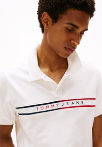 Camiseta polo blanca de algodón con una tapeta de tres botones, que presenta una prominente franja de logo horizontal en rojo, azul marino y blanco a lo largo del pecho.