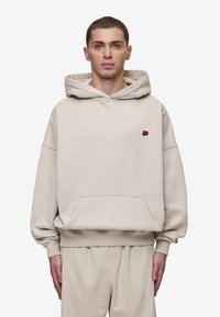Hoodie oversized bege com bolso frontal e punhos canelados. Apresenta um pequeno logotipo rosa no peito. Textura de tecido suave.
