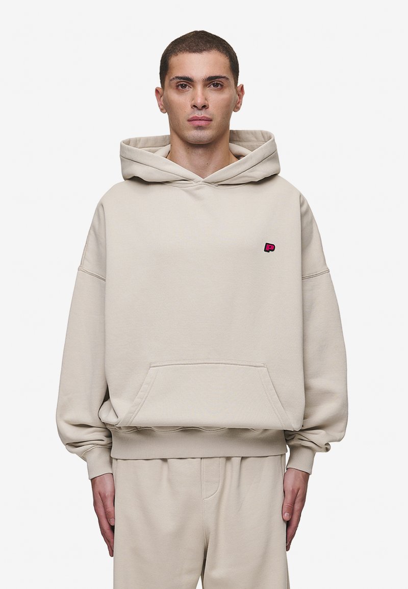 Hoodie oversized bege com bolso frontal e punhos canelados. Apresenta um pequeno logotipo rosa no peito. Textura de tecido suave.