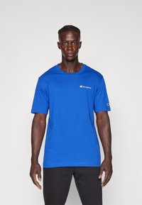 T-shirt en coton bleu avec des manches courtes et un col rond. Présente un petit logo "Champion" sur la poitrine gauche et un logo sur la manche.