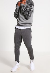 Sudadera gris con mangas negras estampadas, combinada con pantalones grises de rayas finas y zapatillas deportivas blancas. Camiseta blanca lisa debajo.