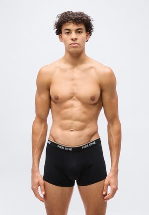 Jeune homme aux cheveux bouclés portant un boxer noir Pier One, debout devant un fond gris clair uni, faisant face à l'avant.