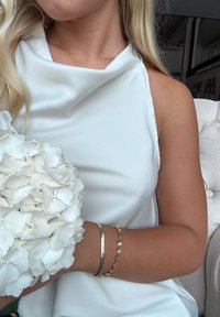 Vit ärmlös klänning med draperad halsringning; håller en bukett av vita hortensior. Två armband: ett enkelt i silver och ett med mönster i guld.