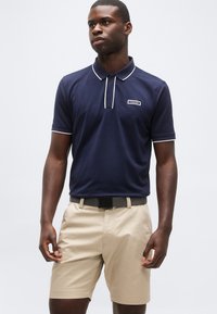 Νεαρός άνδρας που φοράει μπλε πουκάμισο πόλο με λευκή περιγραφή και μπεζ σορτς, στέκεται μπροστά σε ένα απλό φωτεινό φόντο.