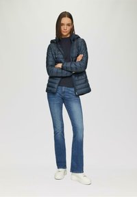 Veste rembourrée à capuche de couleur bleu foncé, associée à un jean ajusté et des baskets à plateforme blanches. La veste se distingue par un design matelassé et des poches zippées.
