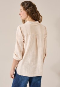 Chemise boutonnée beige clair en tissu texturé et froissé, dotée d'un col et de manches retroussées. Coupe ajustée avec un ourlet légèrement arrondi.