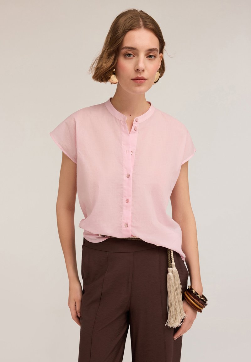 Camicia corta rosa chiaro a maniche corte con colletto alla cinese, chiusura a bottoni e tessuto morbido, abbinata a pantaloni marroni e accessori con un pon-pon.