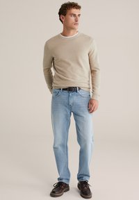 Bež pleteni džemper s okruglim izrezom, u kombinaciji sa svijetloplavim trapericama. Model nosi tamnosmeđe cipele s teksturiranim završetkom.