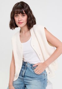 Gilet en polaire de couleur blanc cassé avec des boutons pression, sans manches, en tissu texturé, porté sur un haut blanc et un jean bleu clair.