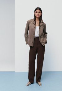 Brun button-up jakke over en hvid top, kombineret med brune bukser og mønstrede spidse sko. Glat stof og afslappet pasform.
