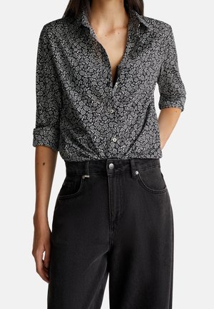 Kobieta ubrana w czarno-białą koszulę w kwiaty z guzikami, z rękawami podwiniętymi do 3/4, wpuszczoną w czarne, wysokowalne dżinsy.