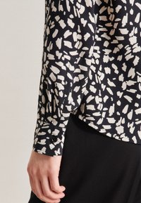 Blusa nera con motivo astratto bianco, maniche lunghe, polsini con bottoni e texture liscia; esposta su un braccio parzialmente visibile.