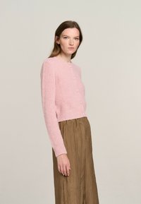 Rosa gestrickte Strickjacke mit langen Ärmeln und Knopfverschlüssen, kombiniert mit einem braunen gefalteten Rock. Weicher Textil und cropped Design.