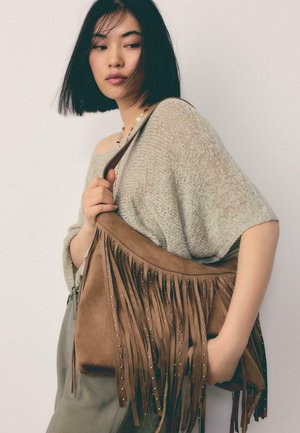 Junge Frau mit glatten schwarzen Haaren, die einen beigen Strickpullover trägt und eine braune Wildlederfransentasche mit Metallnieten über der Schulter trägt.