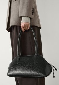 Main tenant un sac à main en cuir noir texturé avec deux poignées, porté avec un blazer taupe et un pantalon foncé sur un fond neutre.