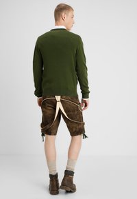 Grüner Pullover, braune Wildledershorts mit kontrastierenden beigen Trägern und Quasten, kombiniert mit braunen Stiefeln und beigen Socken. Lässiges Outfit, Rückansicht.