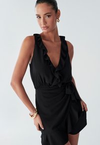 Schwarzes ärmelloses Kleid mit gerafftem Ausschnitt und Taillenschleife, das ein Wickeldesign und einen glatten, fließenden Stoff aufweist. Goldfarbenes Armband am Handgelenk als Akzent.