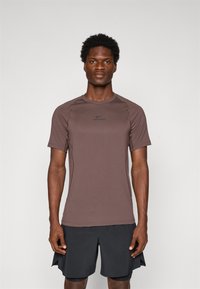 Mann som står vendt mot kameraet iført en brun Nike Pro kortermet skjorte og svarte treningsshorts mot en enkel lys bakgrunn.