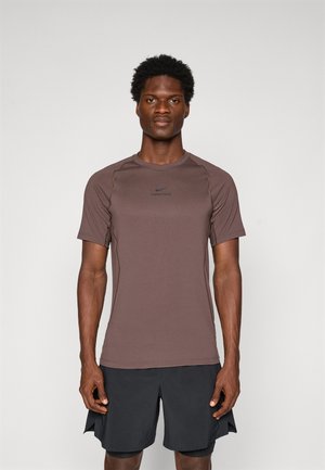 Un homme faisant face, portant un T-shirt à manches courtes Nike Pro marron et un short de sport noir, sur un fond clair et uni.