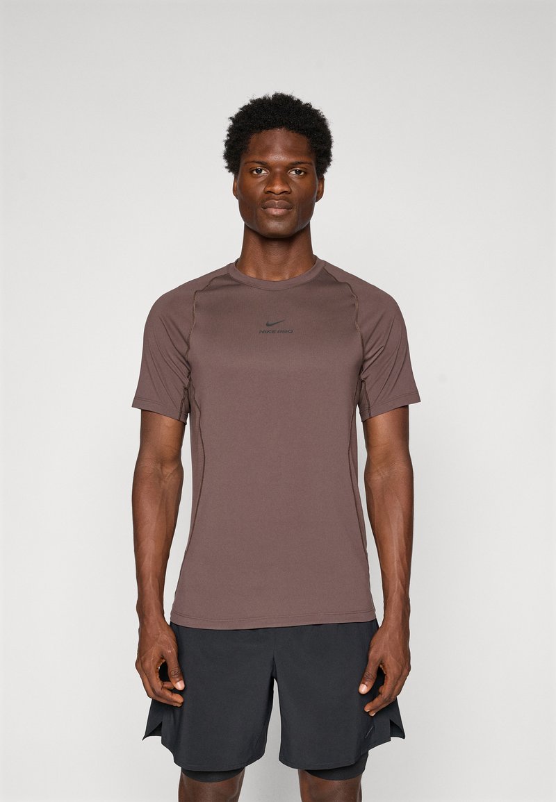 Mann som står vendt mot kameraet iført en brun Nike Pro kortermet skjorte og svarte treningsshorts mot en enkel lys bakgrunn.