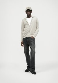 Lys beige zip-up cardigan over en hvid skjorte, parret med mørkegrå jeans og sorte sko. En lys grå cap fuldender outfittet.