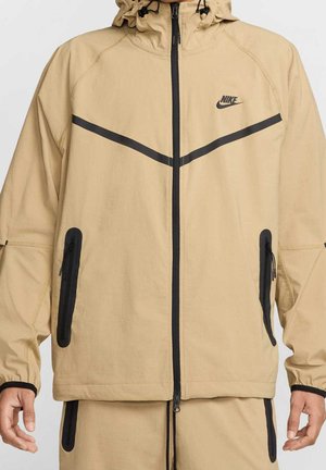 Beige Nike jas met capuchon, zwarte rits, schuine borststrepen, met rits sluitende zijzakken en bijpassende beige broek met zwarte accenten.