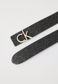 Čierny kožený opasok s embosovaným vzorom loga "CK", doplnený zlatým kovovým sponou so štýlizovaným "CK".