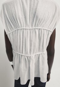 Haut léger en lin blanc avec une texture plissée et froncée, un ourlet incurvé et une taille cintrée. Manches courtes et drapé doux et aéré.