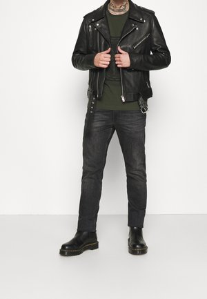 Veste de moto en cuir noir avec des fermetures éclair argentées, t-shirt vert olive, jeans foncés et bottines noires à talons épais, à l'arrière-plan blanc.