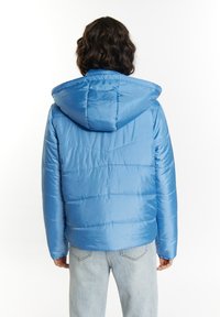 Giacca blu imbottita con cappuccio, caratterizzata da un esterno liscio, texture trapuntata e tasche laterali. Indossata con jeans di denim chiaro.