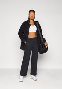 Čierna textúrovaná bunda na zips, biele tričko na crop, čierne nohavice s širokými nohavicami a biele tenisky. Nosiac čiernu crossbody tašku. Neutrálny pozadie.