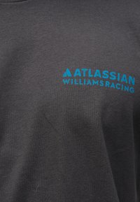 Σκούρο γκρι ύφασμα με φωτεινό μπλε λογότυπο "Atlassian Williams Racing" εκτυπωμένο επάνω του.