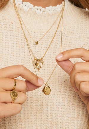 Goudkleurige gelaagde kettingen met een locket-hanger, kleine bedels en parelaccenten. Draagt men over een gemarmerde crèmekleurige trui.