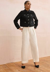 Veste noire courte avec des boutons argentés, associée à un pantalon plissé crème. Des chaussures noires à bout pointu accentuent la tenue. Fond en parquet.