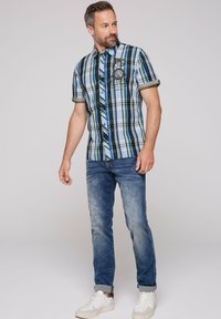 Maglietta a maniche corte a quadri blu, verde e nera con design a bottoni, dotata di taschino sul petto e rifiniture gialle a contrasto. Indossata con jeans in denim e sneakers bianche.