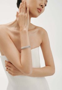 Pulsera de puño de malla plateada con un diseño suave y redondeado. El modelo lleva un vestido blanco sin mangas, destacando la pulsera en su brazo.