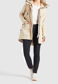 Beżowa wodoodporna parka z regulowanym kapturem, bocznymi kieszeniami i czarnymi elementami; noszona na czarnych spodniach i jasnych sneakersach.