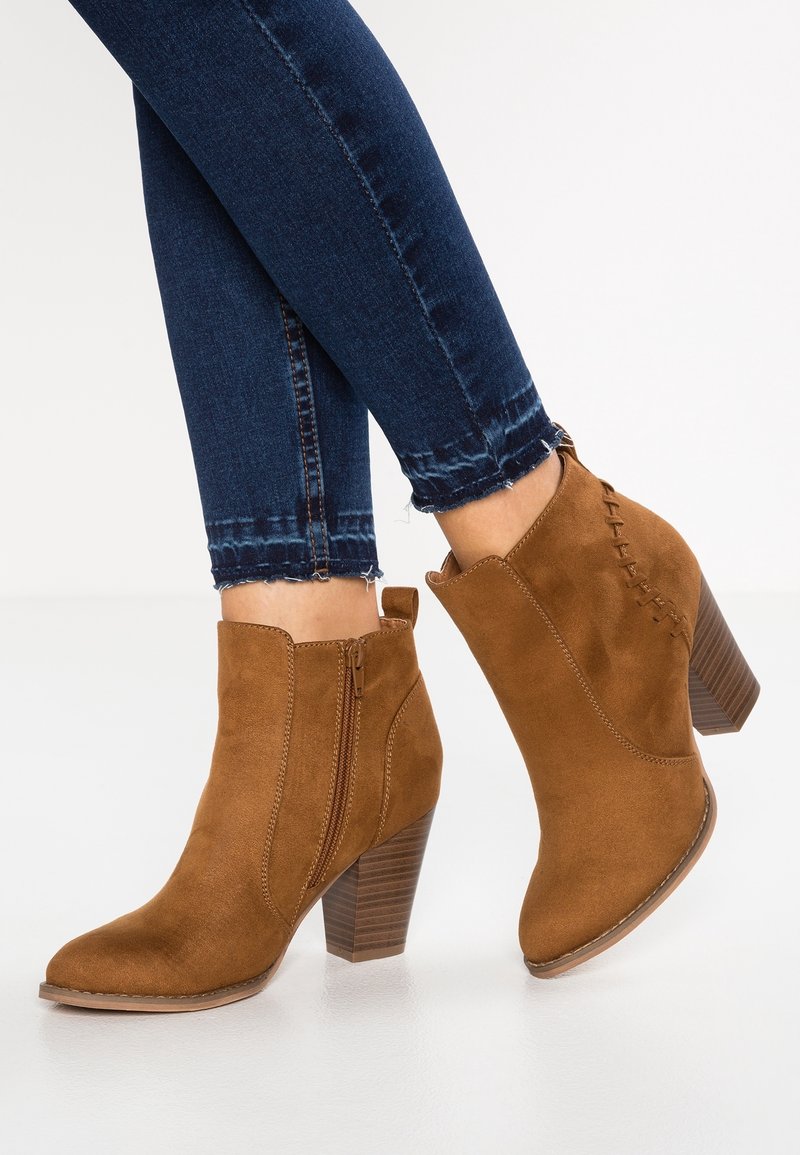 Botines de ante marrón con cremallera lateral, puntera afilada y tacón apilado, que cuentan con costuras decorativas en la parte trasera.