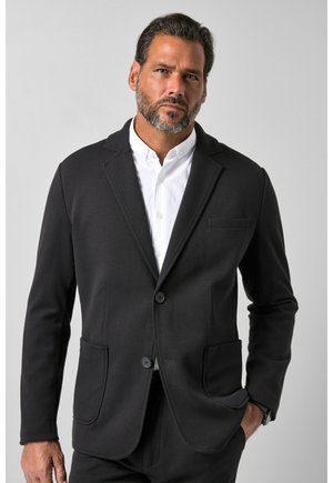 Mann mit Bart, der einen schwarzen Blazer über einem weißen, geknöpften Hemd trägt, steht mit der Hand in der Tasche vor einem einfarbigen hellen Hintergrund.