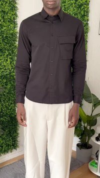 Man in een zwart button-up overhemd met een grote borstzak en witte broek, staande binnen bij groene planten en een gestructureerde muur.