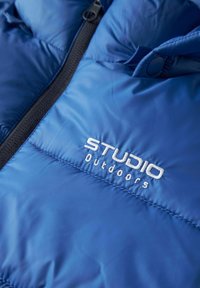 Veste matelassée bleue avec fermeture éclair noire et texte blanc brodé "STUDIO outdoors" sur la poitrine à l'avant.