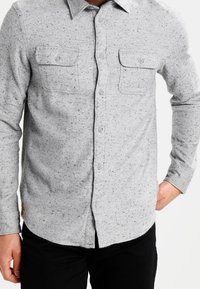 Světlé šedé flanelové košile s texturou posetou tečkami, s knoflíkovou légou, dvěma náprsními kapsami a zaobleným lemem. Viditelné černé kalhoty.