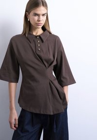 Mlada ženska z ravnimi lasmi, ki nosi rjavo polosrajco z gumbi in naborki ter široke temno modre hlače, stoji ob beli steni.