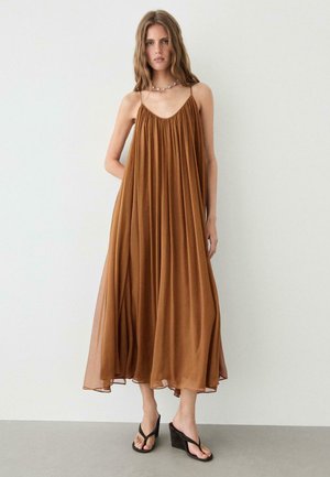 Femme portant une robe midi marron ample sans manches avec fines bretelles et des sandales compensées noires, debout contre un mur blanc uni.